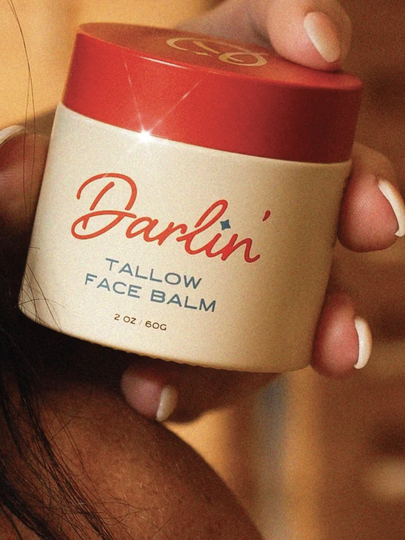 Person holding a container of "Darlin' Tallow Face Balm" with a red lid and beige label.