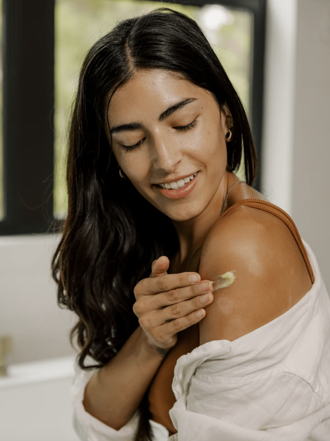 Woman applying lotion to her shoulder, wearing a white shirt with a brown strap visible, and smiling slightly.