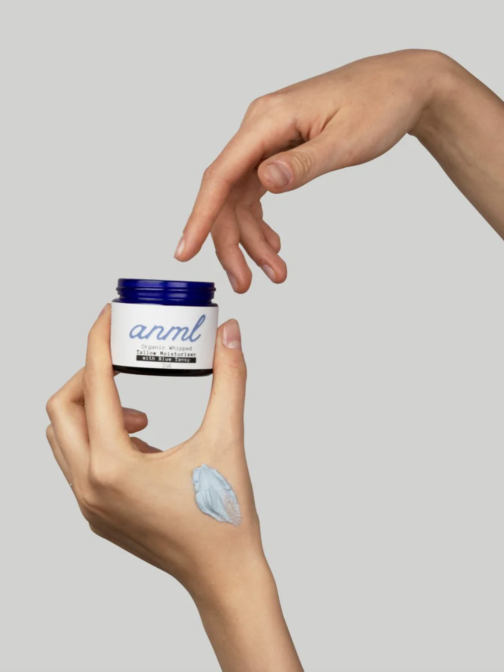 A person's hands holding a jar of skincare cream labeled "anml," with a small amount of cream applied to the back of one hand, against a plain background.