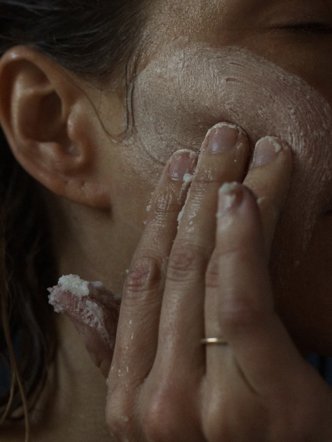 A person applies a creamy facial mask to their cheek with their fingers.