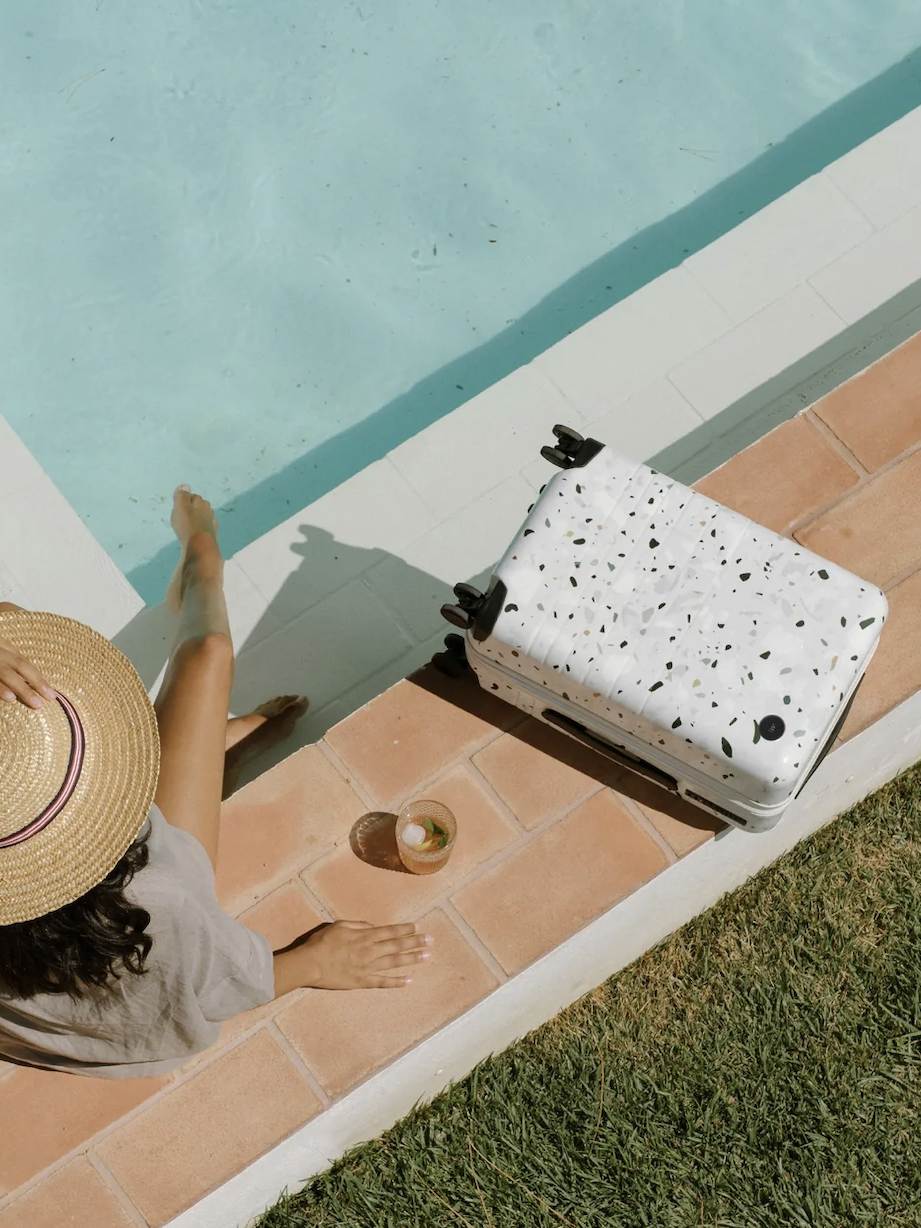 Person sitting at the edge of a pool, wearing a straw hat and dipping feet in water. A white terrazzo-patterned suitcase and a drink are next to them on the tile.
