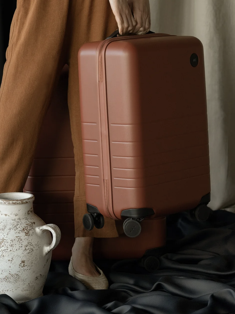 Person holding a brown suitcase, standing next to another identical suitcase on the floor. A decorative vase is visible in the foreground.