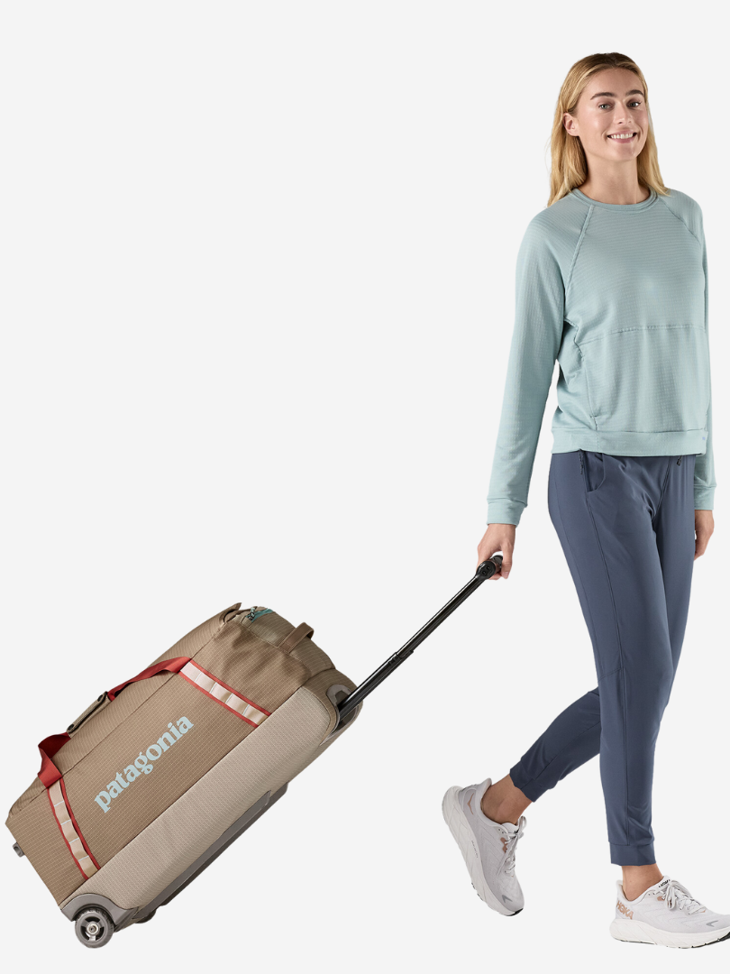 A person in a light blue sweater and blue pants pulls a beige rolling suitcase.