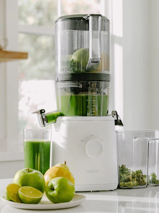 Juicer on a kitchen counter with a glass of green juice, a bowl of lemons and apples, and pulp container.