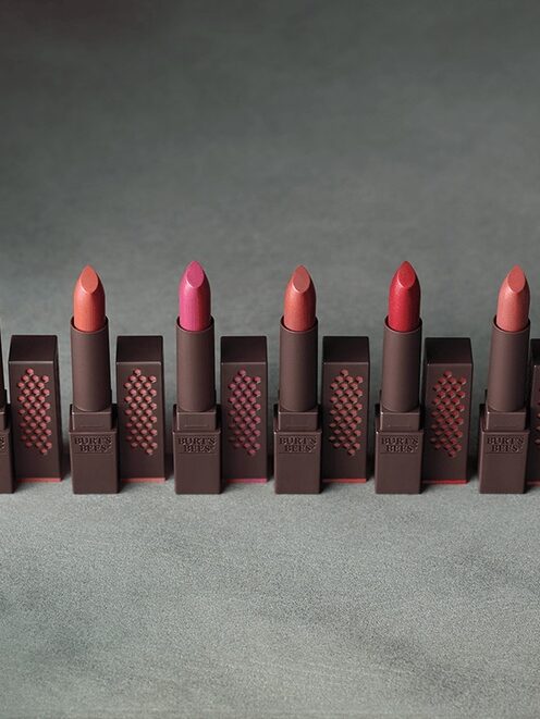Six tubes of lipstick in varying shades of red and pink are arranged in a row on a textured surface.