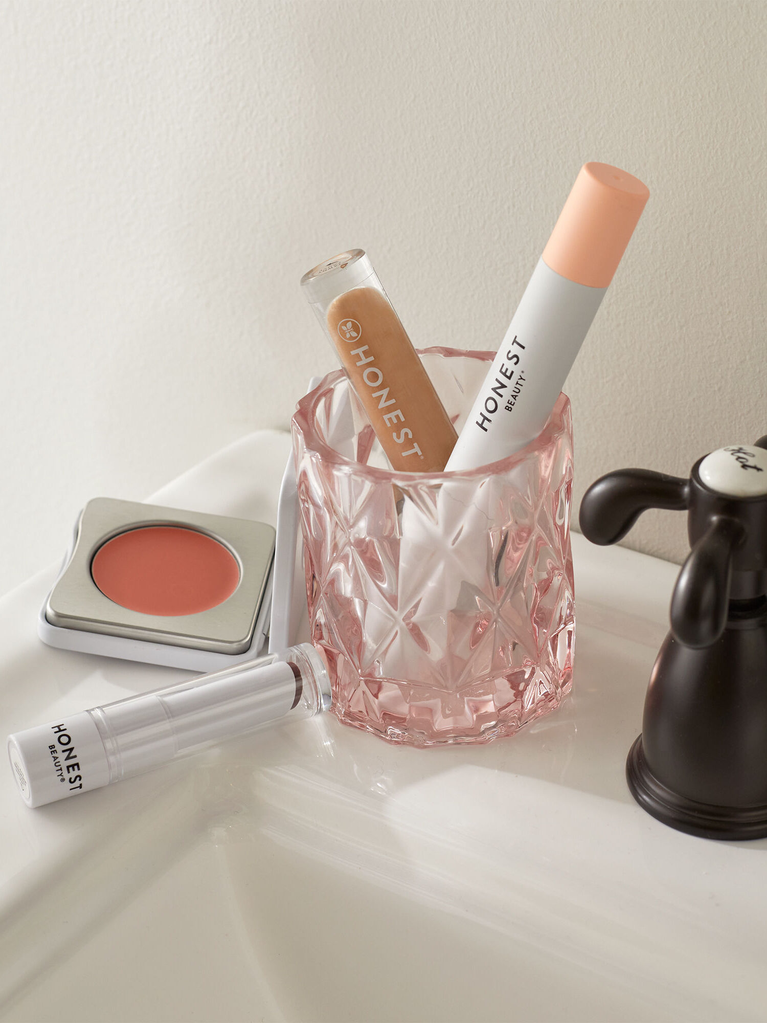 Cosmetic products including lip gloss, concealer, and a compact blush placed on a bathroom sink next to a pink glass.