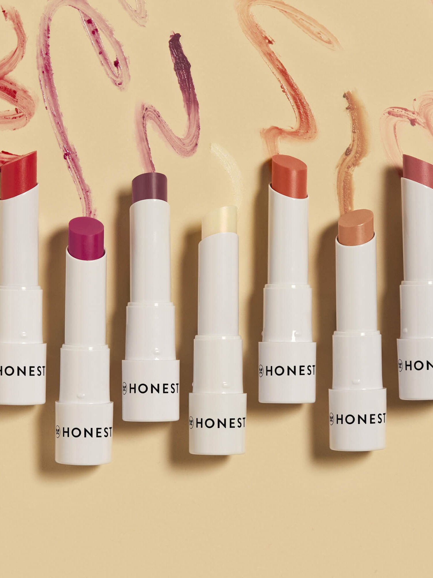 Seven Honest Beauty lipsticks in various shades arranged in a row with smudged samples above on a beige background.