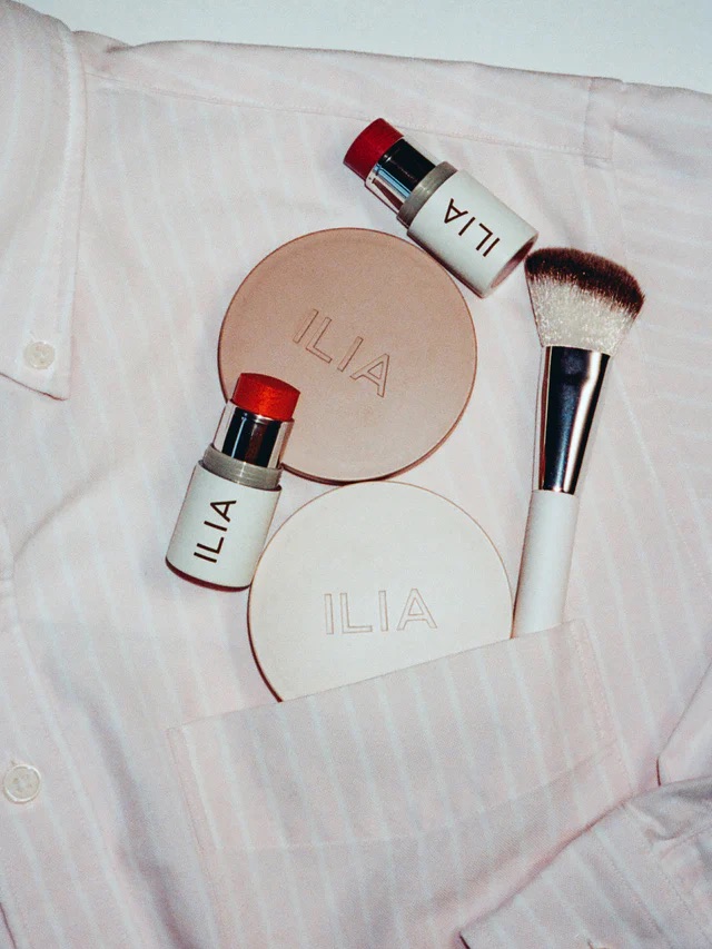 Makeup items labeled ILIA, including two round compacts, two lipsticks, and a brush, are placed on a pink striped shirt.