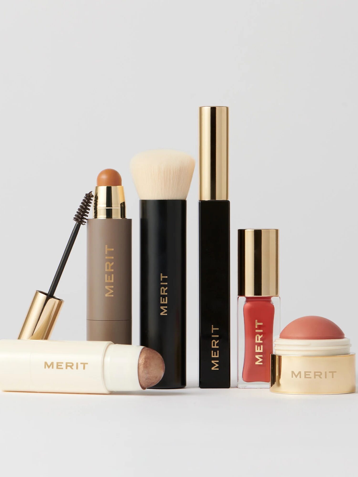 Assorted Merit brand makeup products including a brush, mascara, lipstick, and sticks, all with gold accents, arranged upright on a light background.