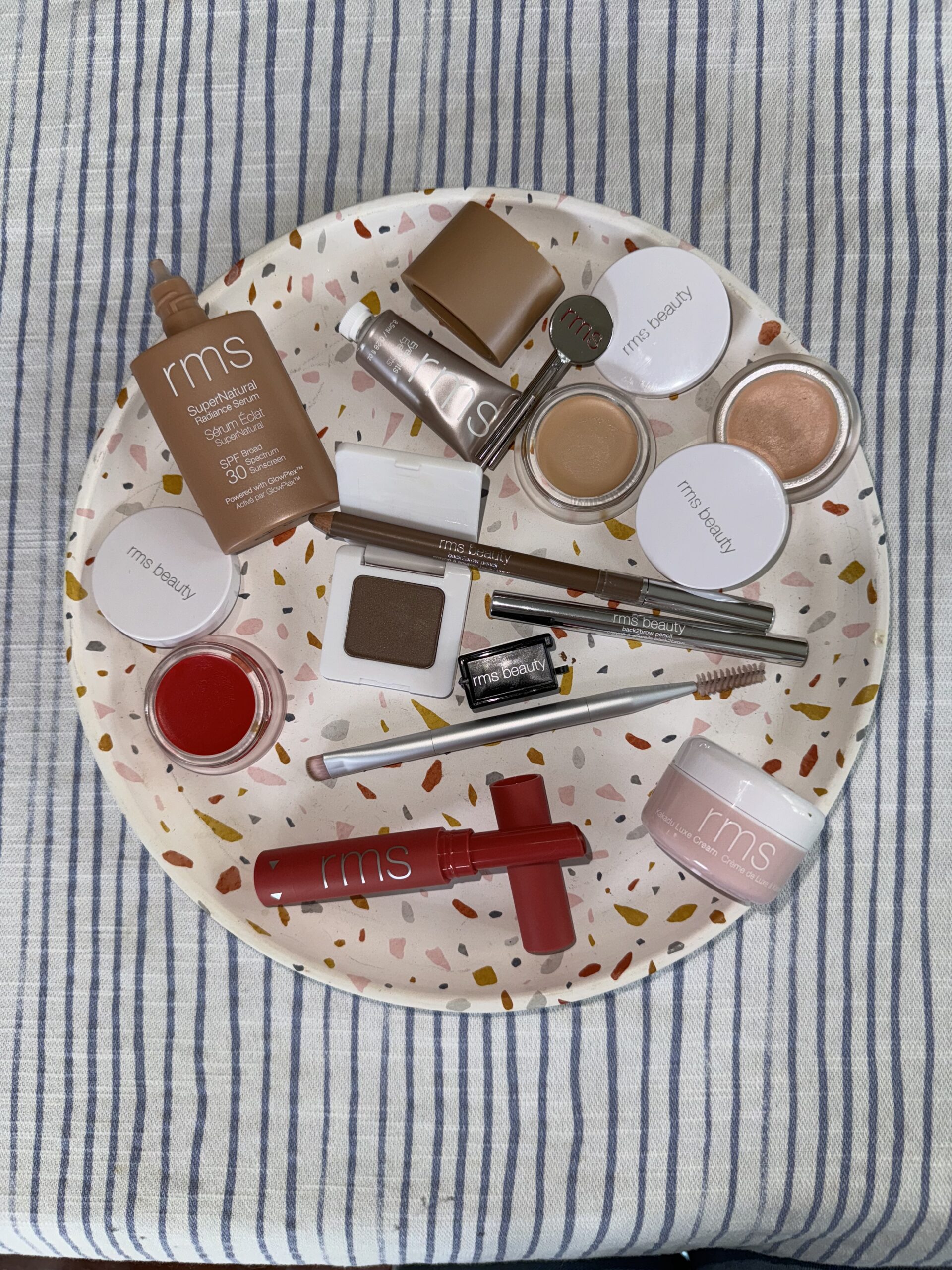 A variety of RMS Beauty makeup products arranged on a speckled circular tray placed on a striped surface.