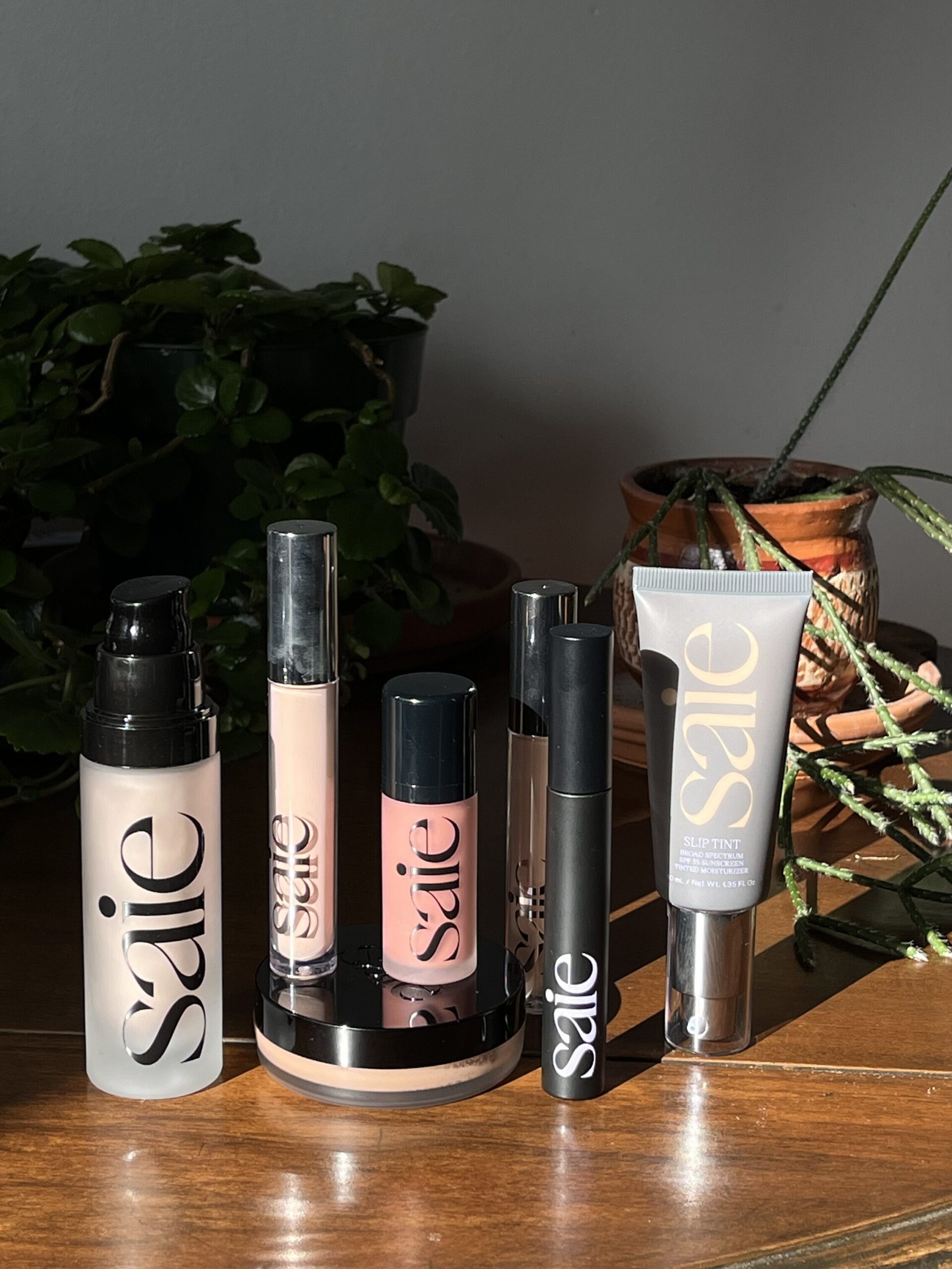 Collection of Saie cosmetic products arranged on a wooden surface, including bottles, tubes, and a compact, with plants in the background.