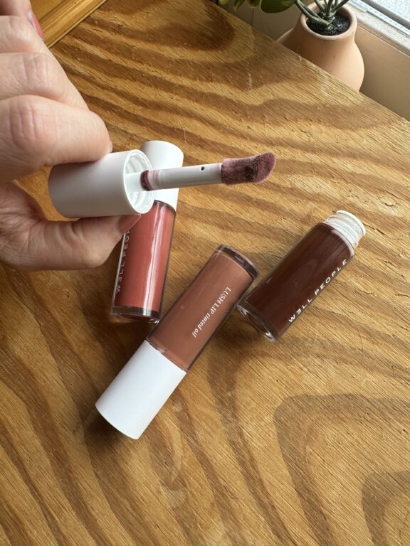 Three tubes of liquid lipstick are on a wooden surface. One tube is open, showing the applicator.
