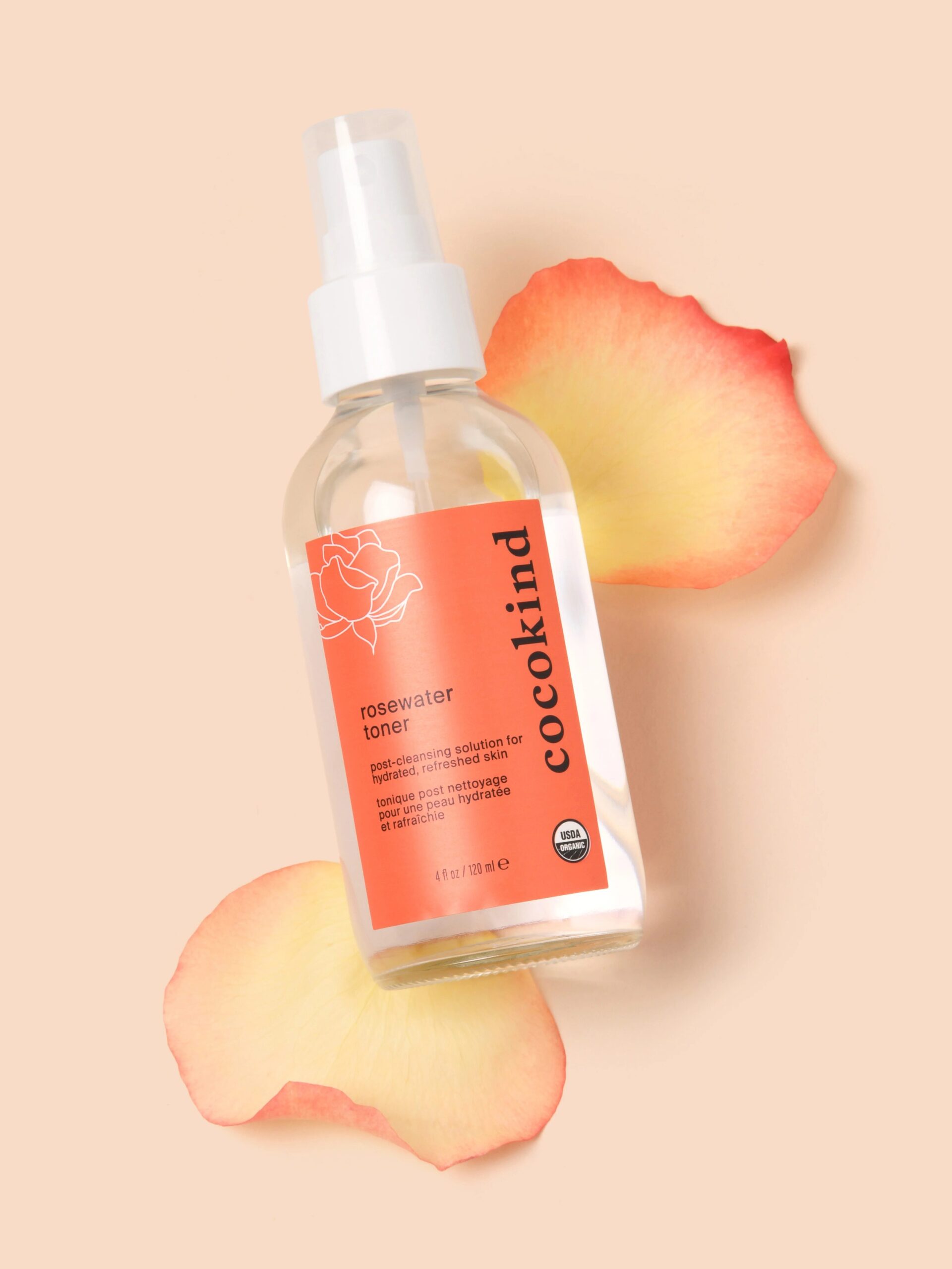 A bottle of Cocokind rosewater toner with an orange label is placed on a beige background, surrounded by three rose petals.