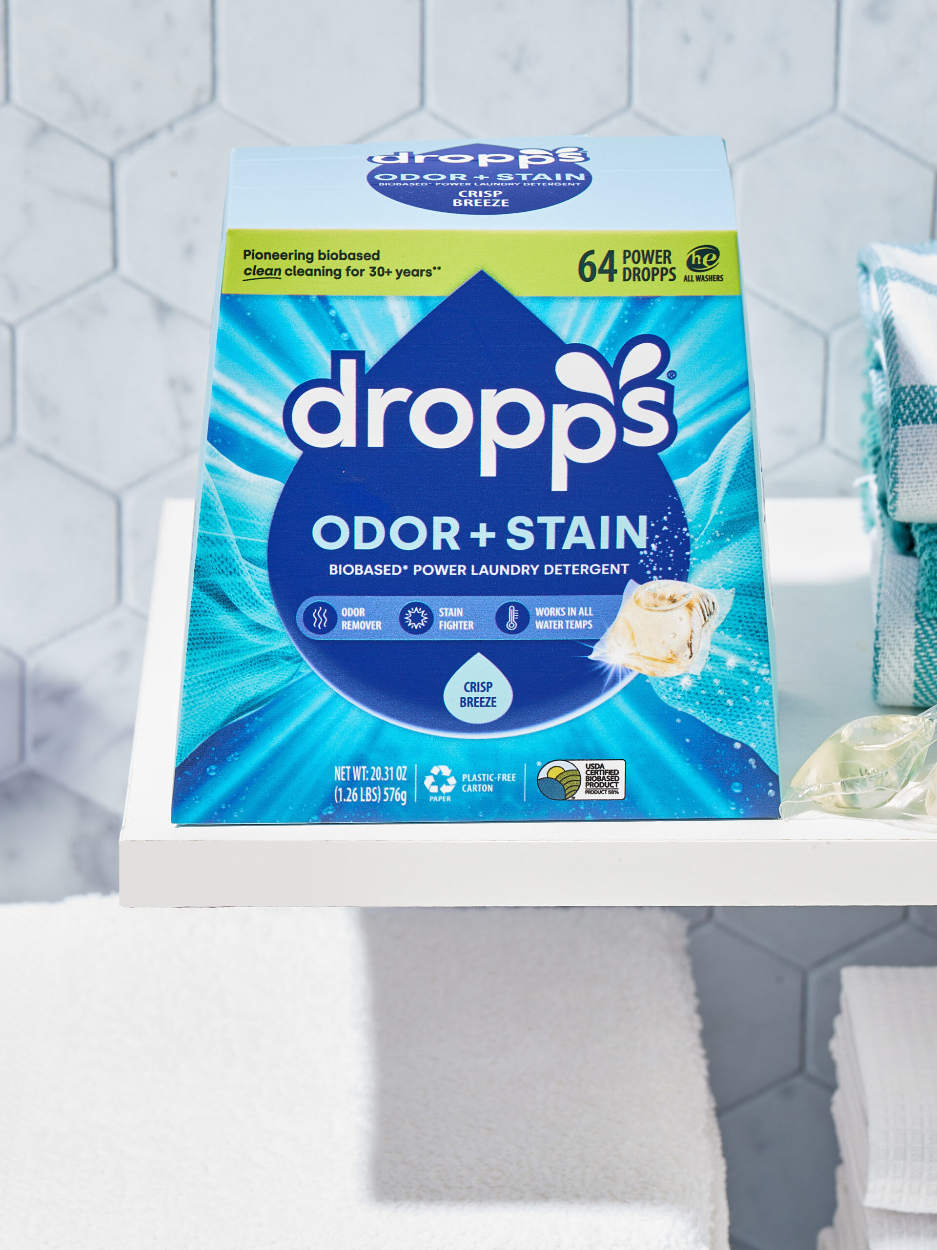 Box of Dropps Odor + Stain laundry detergent pods on a shelf next to two pods, a green plant, folded towels, and a hexagonal tiled wall background.