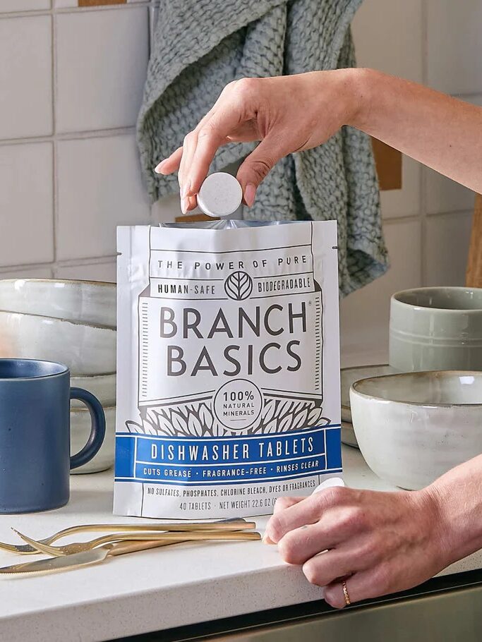 A person holds a dishwasher tablet over an open bag of Branch Basics dishwasher tablets on a kitchen counter with mugs and bowls.