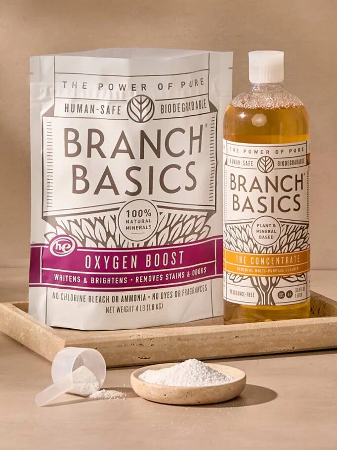 Branch Basics cleaning products displayed; includes a bag labeled "Oxygen Boost," a bottle labeled "The Concentrate," a measuring scoop, and a small pile of white powder on a wooden tray.