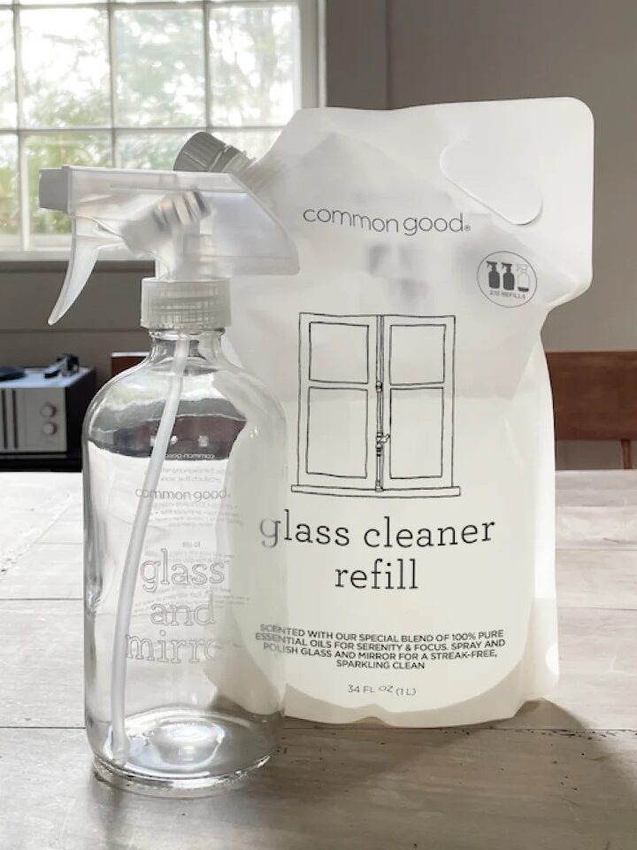 A bottle of glass cleaner and a refill pack on a table, with a window and some furniture in the background.