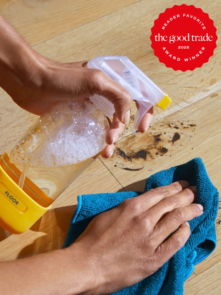 Hands cleaning a wooden floor with a spray bottle and a blue cloth, removing a dark stain. Red award badge in upper right corner.
