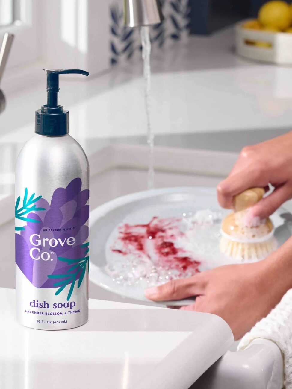 Person washing a plate with a brush in a kitchen sink. A bottle of Grove Co. lavender blossom & thyme dish soap is placed nearby. Lemons are visible in the background.