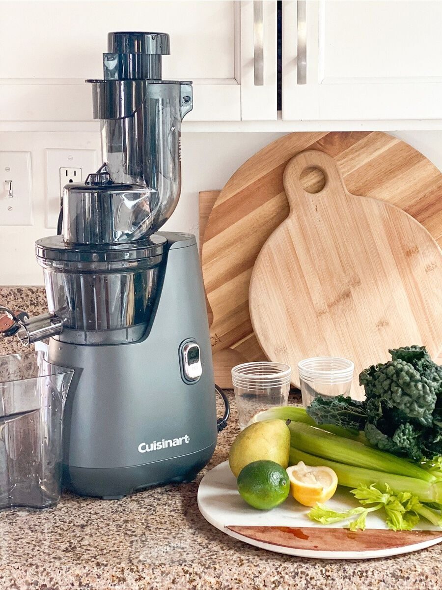 A juicer on a kitchen counter next to leafy greens, celery, a lemon, a lime, and a pear on a cutting board. Two wooden boards are in the background.