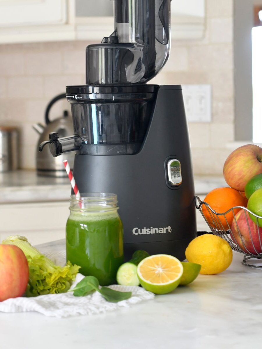 Black juicer on a kitchen counter with a jar of green juice, surrounded by apples, celery, lemon, lime, and oranges.