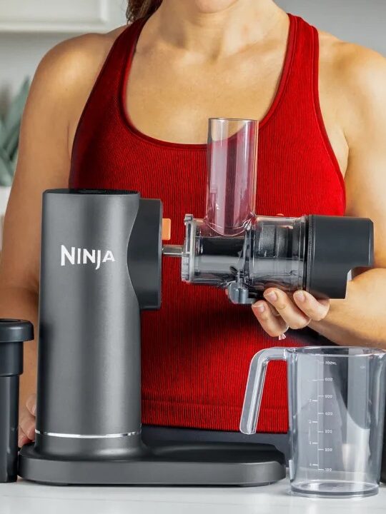 Person in a red tank top assembling a Ninja kitchen appliance on a counter, with additional components and a measuring cup displayed.