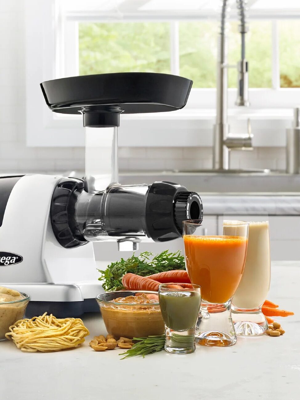 A white Omega cold press juicer on a kitchen counter with various foods including pasta, juice, and nuts. Text highlights its multifunctionality for sorbet, nut butter, pasta, and more.