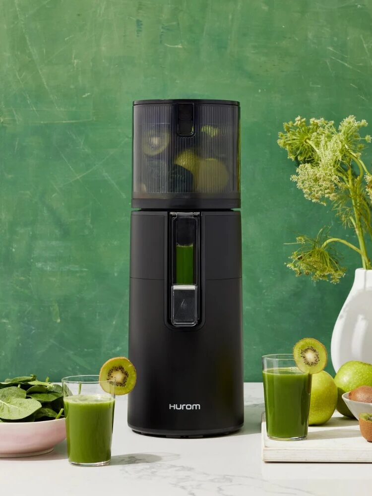 A black juicer with green juice inside is on a countertop. Two glasses of green juice, spinach leaves, kiwi, and apples are next to it. A vase with greenery is in the background.