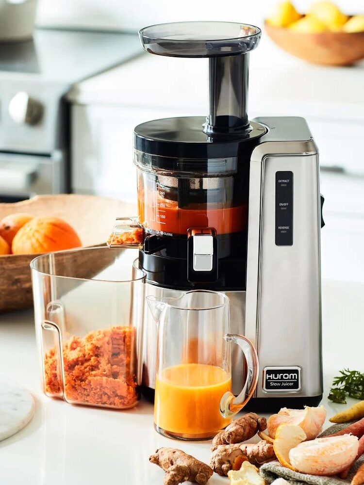 A silver juicer on a kitchen counter with an orange drink in a glass. A wooden bowl of oranges and a cutting board with ginger and other produce are in the background.