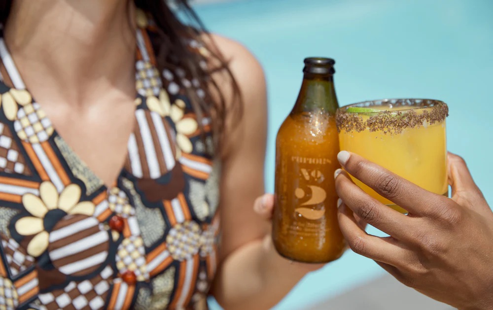 Two people by a pool holding drinks: one with a bottle of kombucha and the other with a glass of yellow beverage with a chili salt rim.