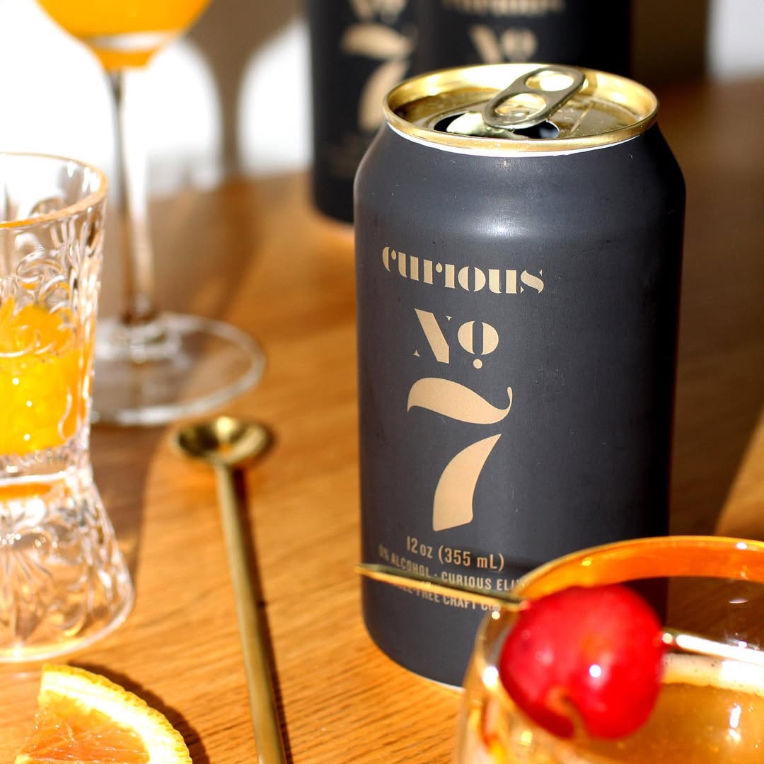 A black can labeled "Curious No. 7" sits on a wooden table surrounded by cocktail glasses, garnished with fruit.