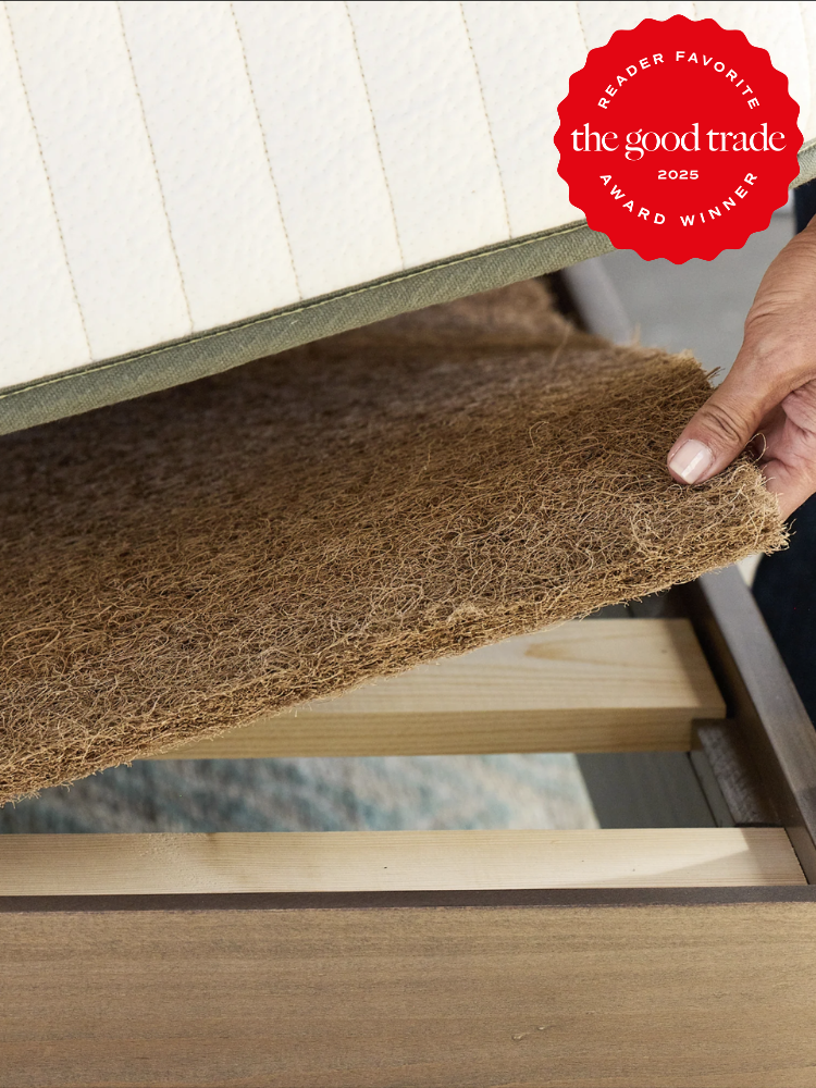 A hand lifting a layer of natural fibers in a wooden bed frame. A red badge reads "Reader Favorite, The Good Trade, Award Winner 2025.