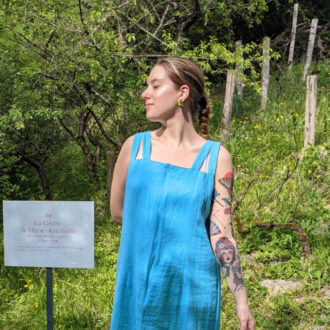 A person in a blue dress stands outdoors near a grassy area with trees. There's a sign nearby in French. The person has a tattoo on one arm and is looking to the side.