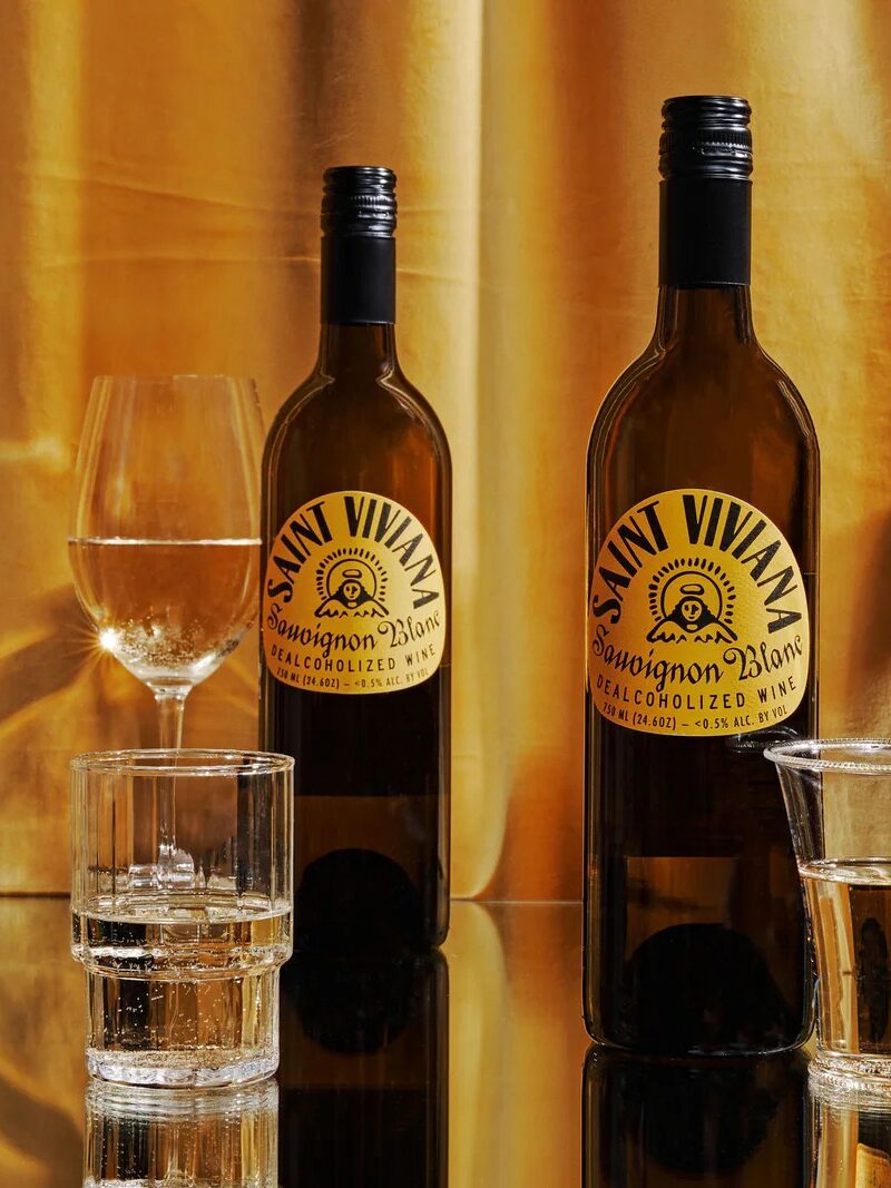 Three Saint Vivant Sauvignon Blanc wine bottles and several glasses filled with white wine on a reflective surface in front of a gold curtain.