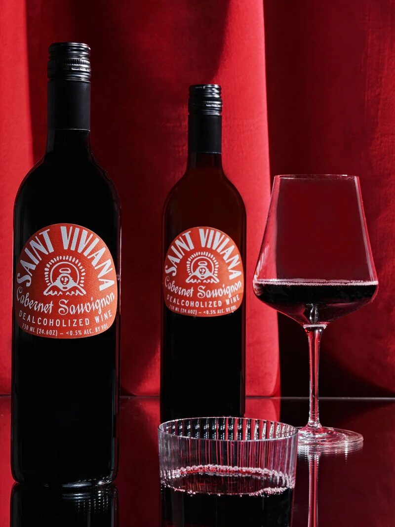 Three bottles of non-alcoholic red wine labeled "Saint Vivant" stand on a reflective surface, accompanied by two filled wine glasses, against a red curtain backdrop.