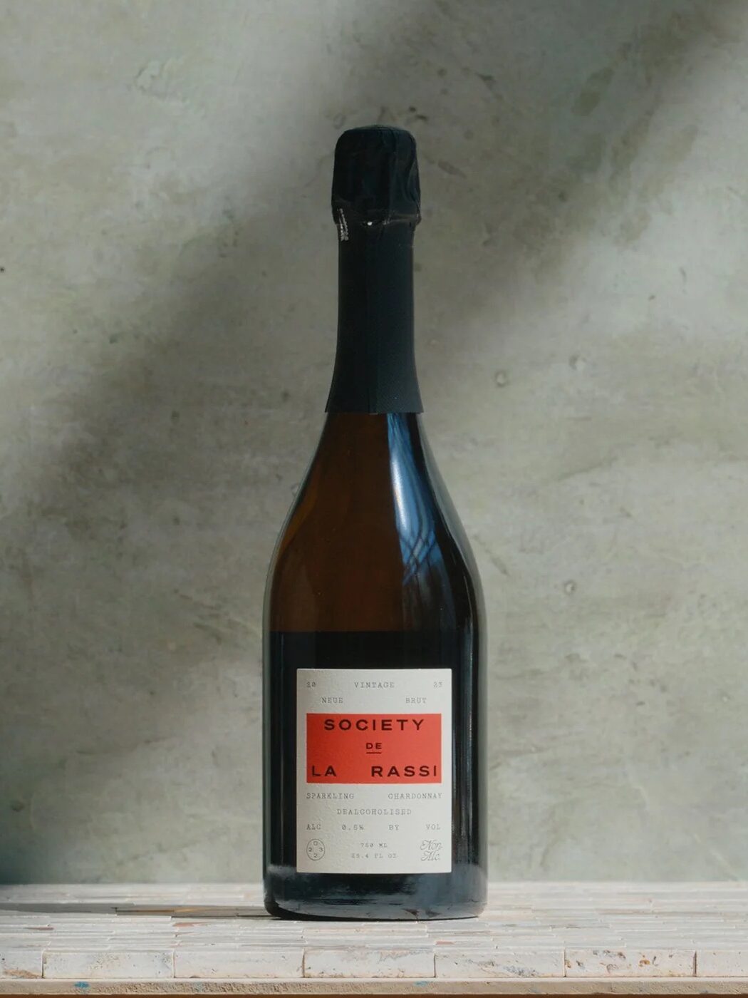 Bottle of sparkling wine with red label on a light textured surface, against a gray concrete wall backdrop.