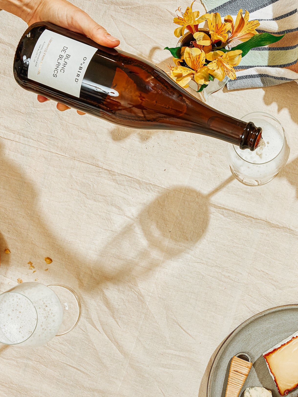 Hand pouring champagne into a glass on a table with bread, cheese, walnuts, and a bouquet of yellow flowers.