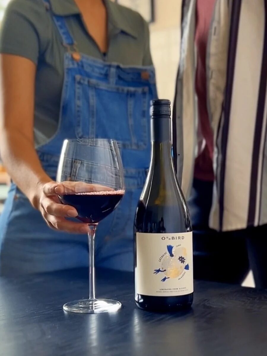 Two people stand by a table with a bottle of wine and a wine glass. One person is holding the glass. They are indoors with large windows in the background.