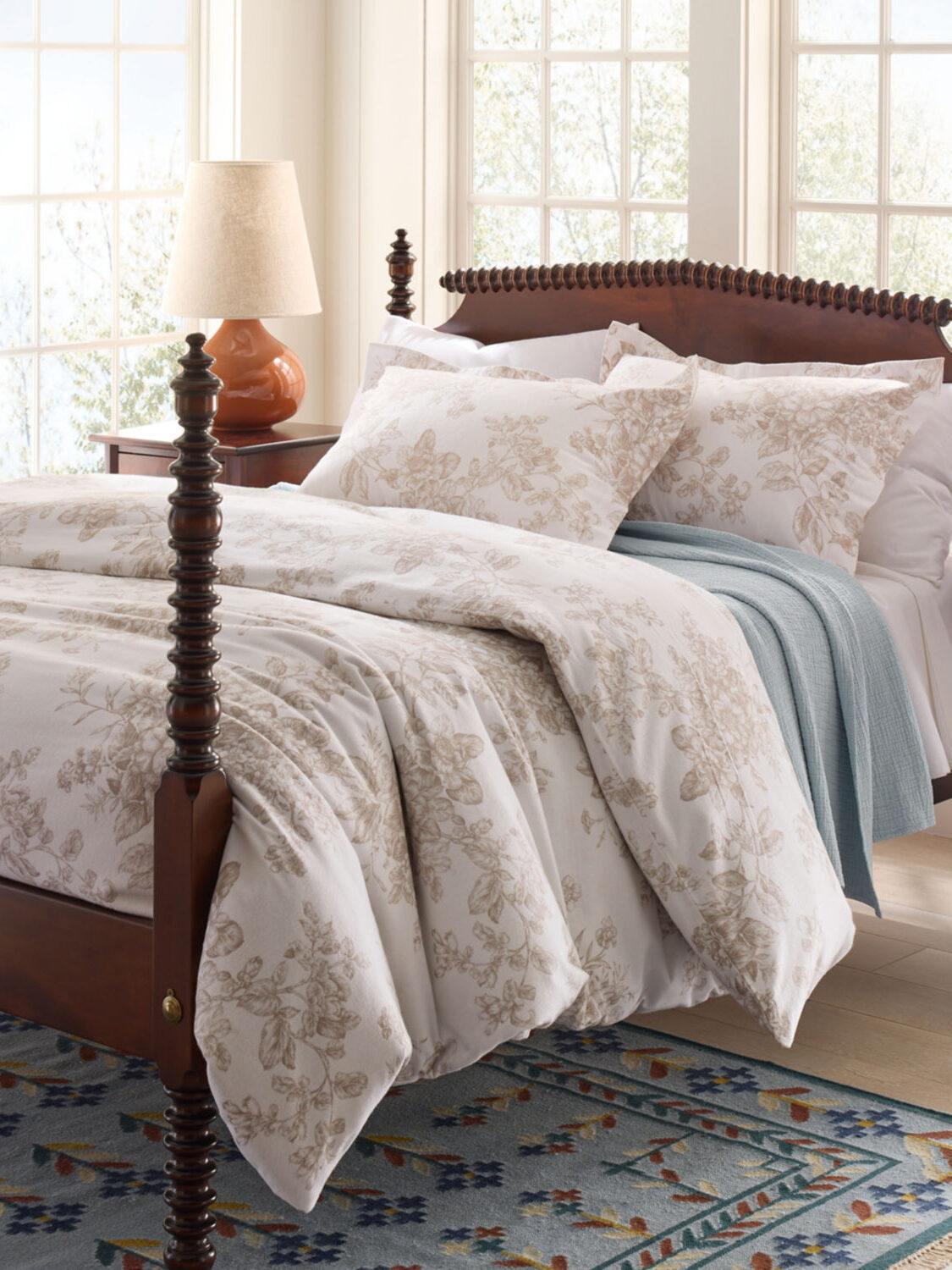 A neatly made bed with floral-patterned bedding, a wooden four-poster frame, and a nearby nightstand with a lamp. Large windows in the background allow natural light to enter the room.
