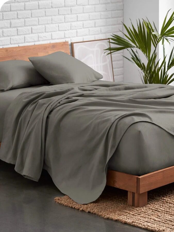 A neatly made bed with gray bedding sits on a wooden frame, next to a white brick wall and window. A green plant is placed by the bed, and a textured rug lies on the floor.