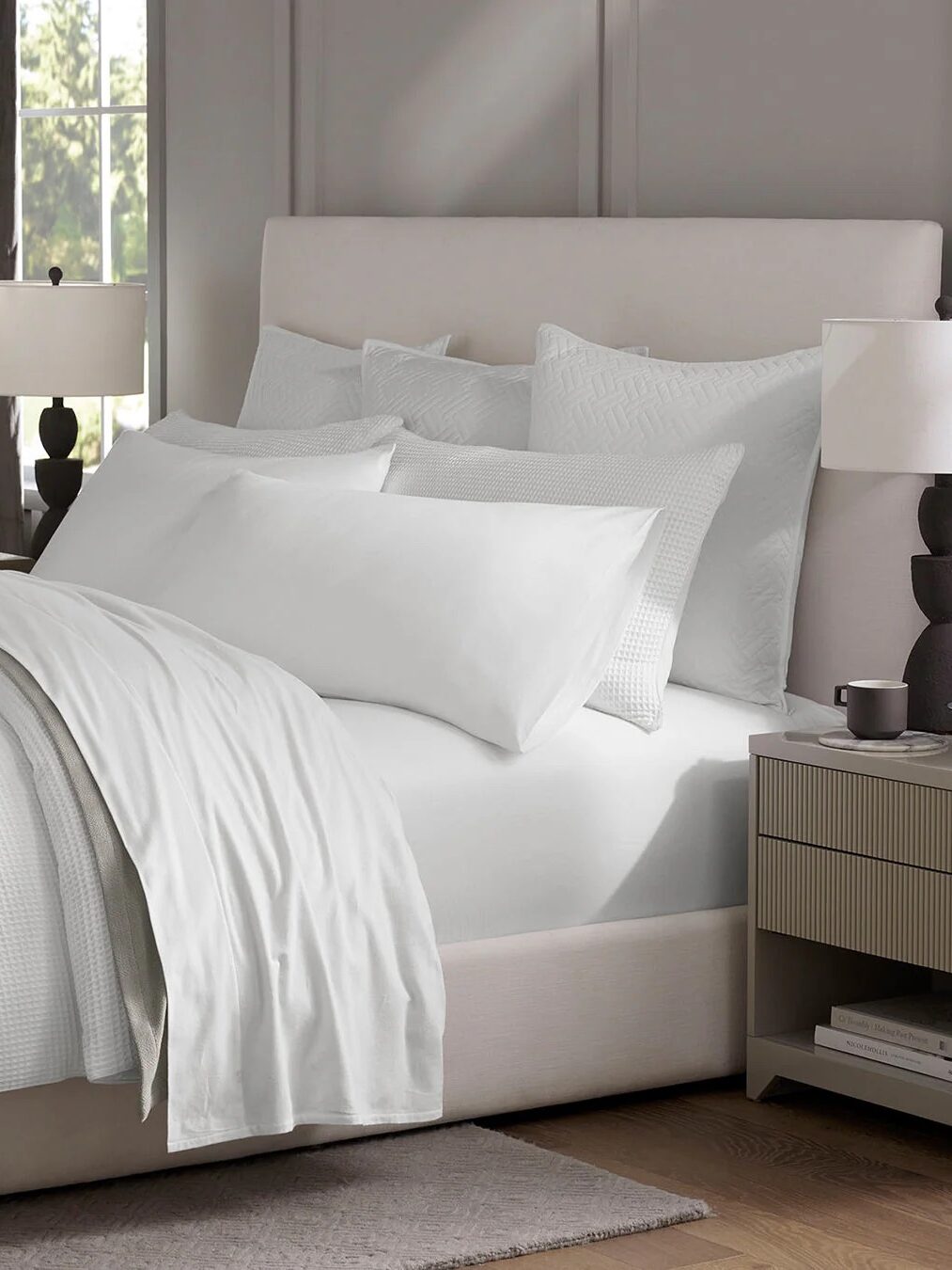 Modern bedroom with a neatly made bed featuring white linens and multiple pillows. A beige headboard, bedside tables with lamps, and a window with curtains complete the setting.