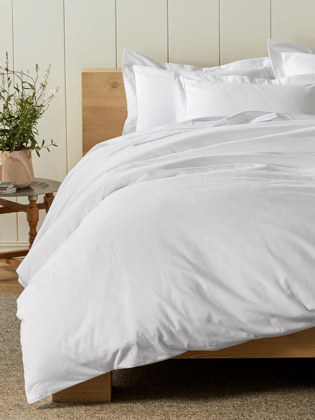 A neatly made bed with white bedding, situated in a room with a wooden headboard and a small side table holding a plant.