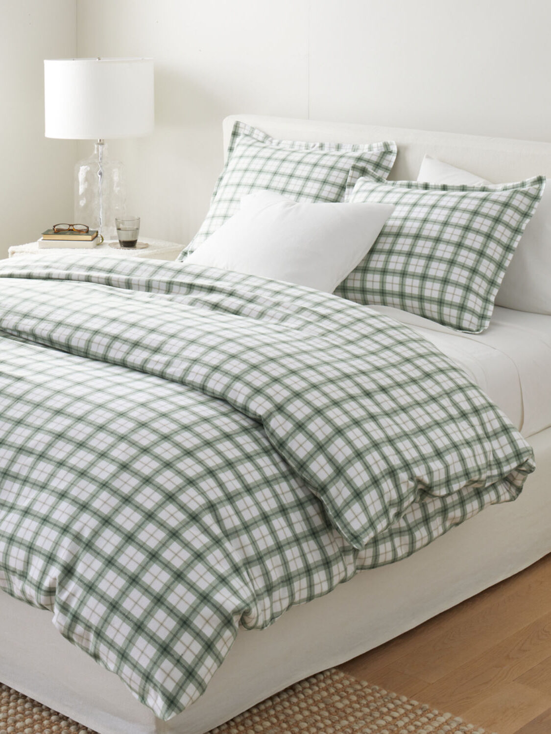 A neatly made bed with green and white plaid bedding, two tartan pillows, and two plain white pillows. A wooden nightstand holds a lamp and books.
