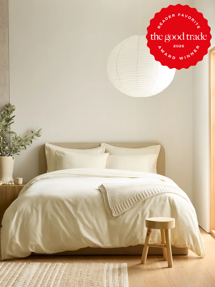 Minimalist bedroom with a light wood bed, white bedding, a small wooden stool, a plant on a side table, and a hanging round paper lantern. A "The Good Trade" award badge is in the top corner.