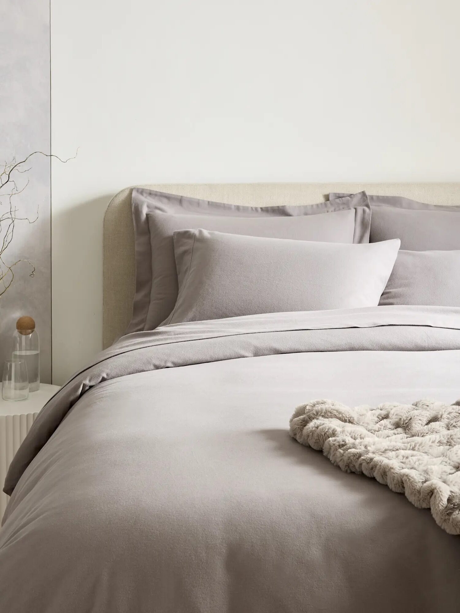 A neatly made bed with gray bedding and pillows. A small side table holds a glass bottle, a wooden object, and decorative twigs. A textured blanket is draped at the foot of the bed.