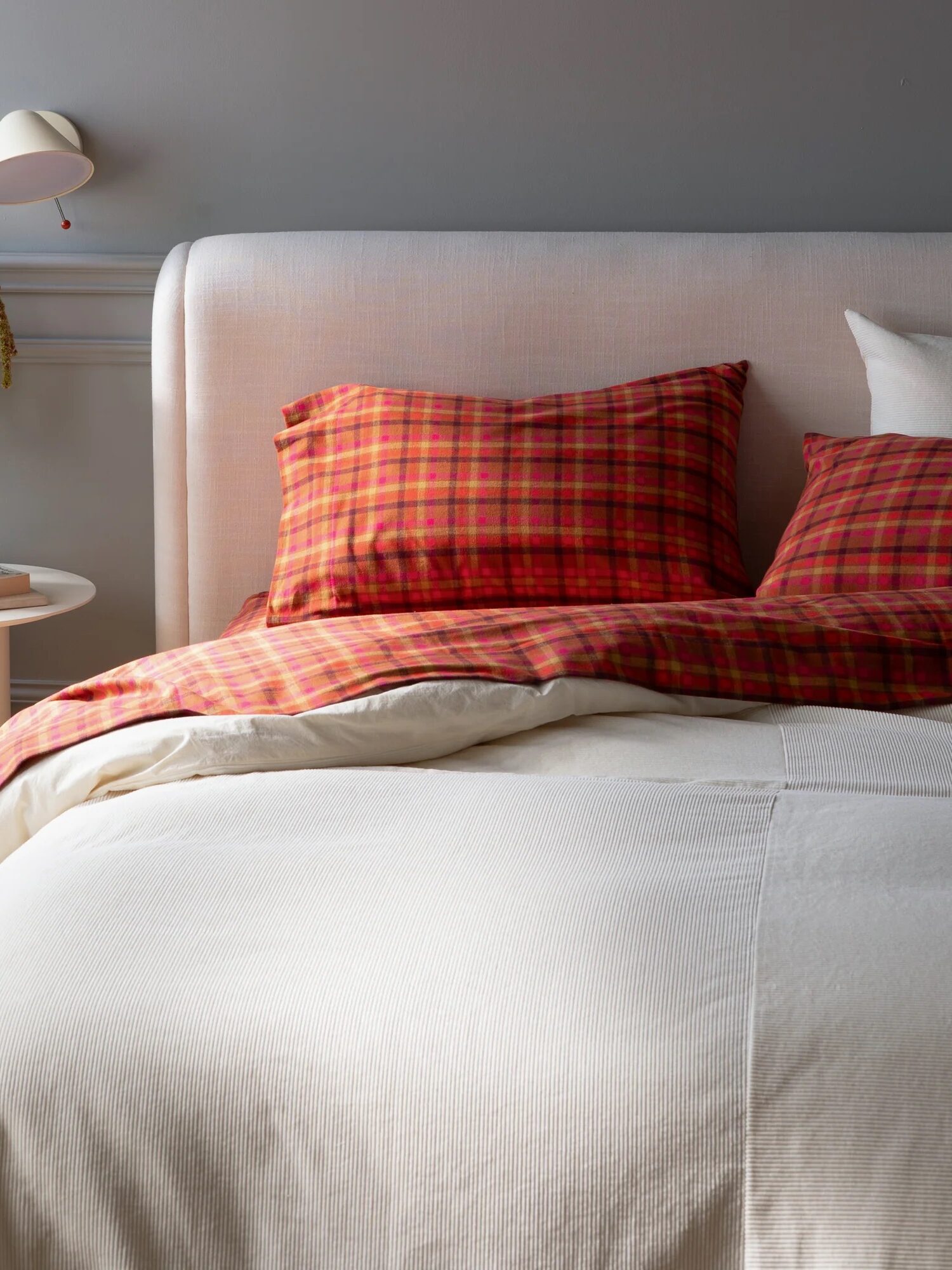 Bedroom scene with a neatly made bed featuring red plaid pillows and a white comforter. A round table beside the bed holds a green plant and a small cup under a wall-mounted lamp.