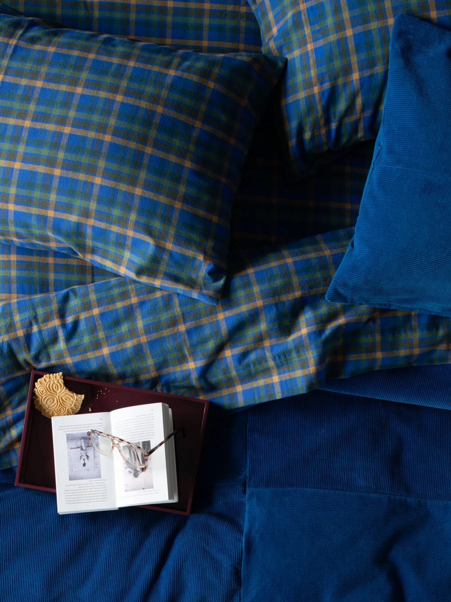 A bed with blue plaid bedding, a tray holding an open book, a pair of glasses, and a snack resting on the bedspread.