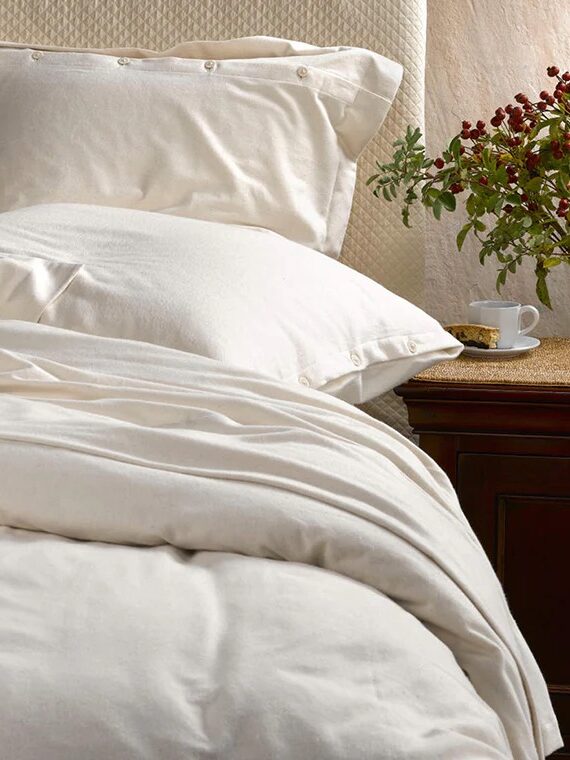 A neatly made bed with white sheets and pillows beside a wooden nightstand with a plant and a white teacup.