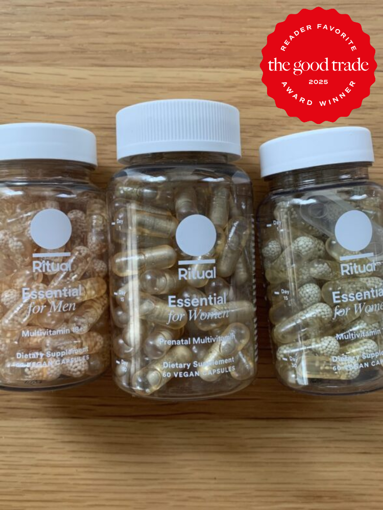 Three bottles of Ritual multivitamins labeled "Essential for Men" and "Essential for Women" and a prenatal vitamin bottle on a wooden surface. A red badge reads "Reader Favorite, the good trade, 2023 Award Winner.
