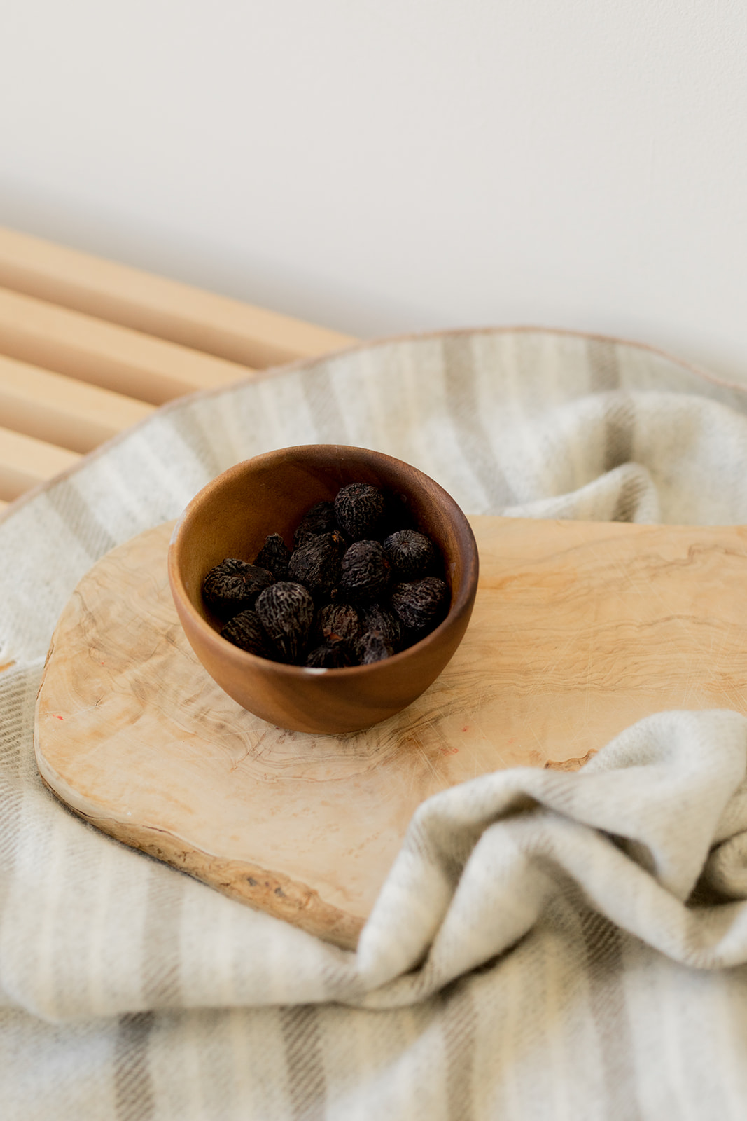 A wooden bowl filled with small, dark fruit sits on a wooden board atop a striped cloth.